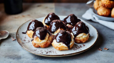 Light cream profiteroles filled with whipped cream and dipped in silky dark chocolate sauce on a plate.