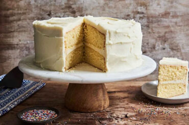 A vanilla cake on a cake stand with one slice cut out of it.