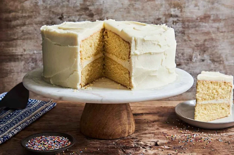 A vanilla cake on a cake stand with one slice cut out of it.