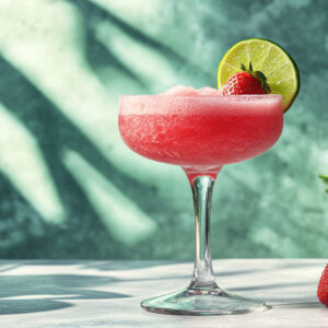 A photo showing a drink made from a strawberry daiquiri recipe. The strawberry daiquiri is in a cocktail glass with a lime and strawberry on the side of the glass.